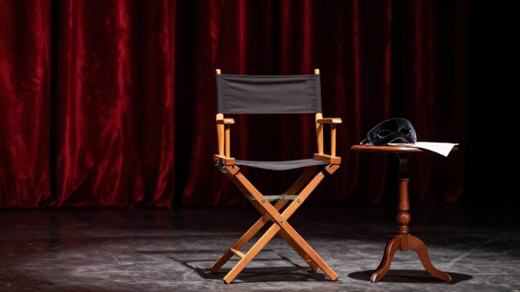 Silla de director de madera junto a una máscara de terciopelo y un guion sobre una mesa, en un escenario de teatro con cortinas rojas de fondo.