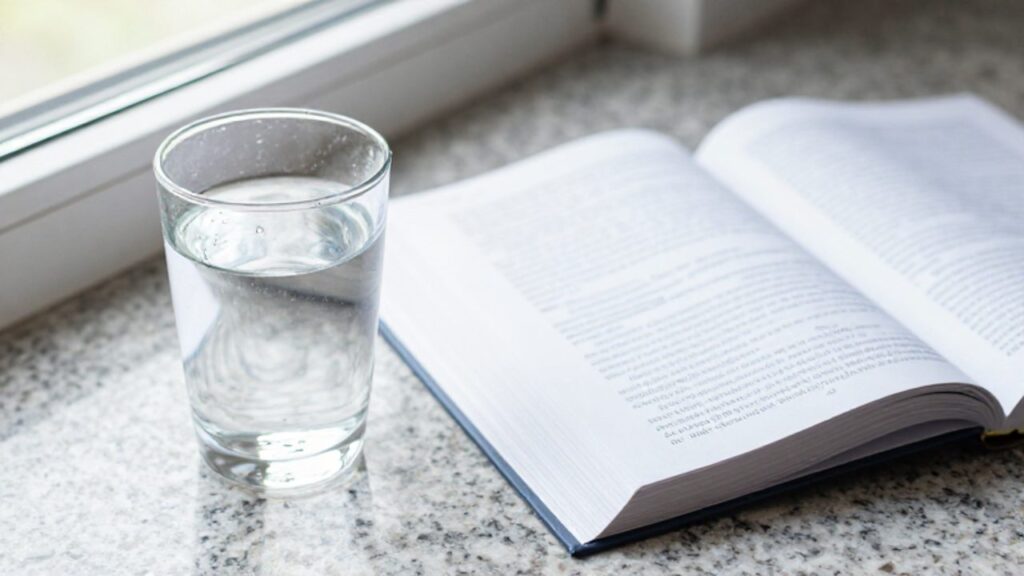 Un vaso de agua con gotas de condensación junto a un libro de texto abierto sobre una mesa de granito.