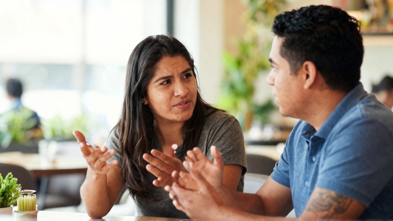 Dos personas latinoamericanas conversan con gestos expresivos sentadas en una cafetería luminosa.